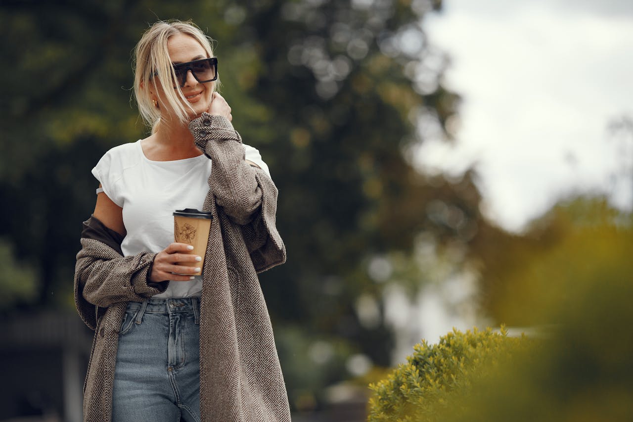 A stylish young woman enjoys a coffee while walking in a sunny, leafy park.